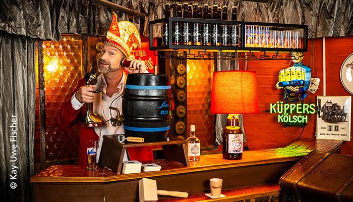 Volker Weininger, der Sitzungspräsident.  ist in seiner Kellerbar mit Narrenkappe, anachronistischem Telefon und Kölsch Werbung zu sehen.